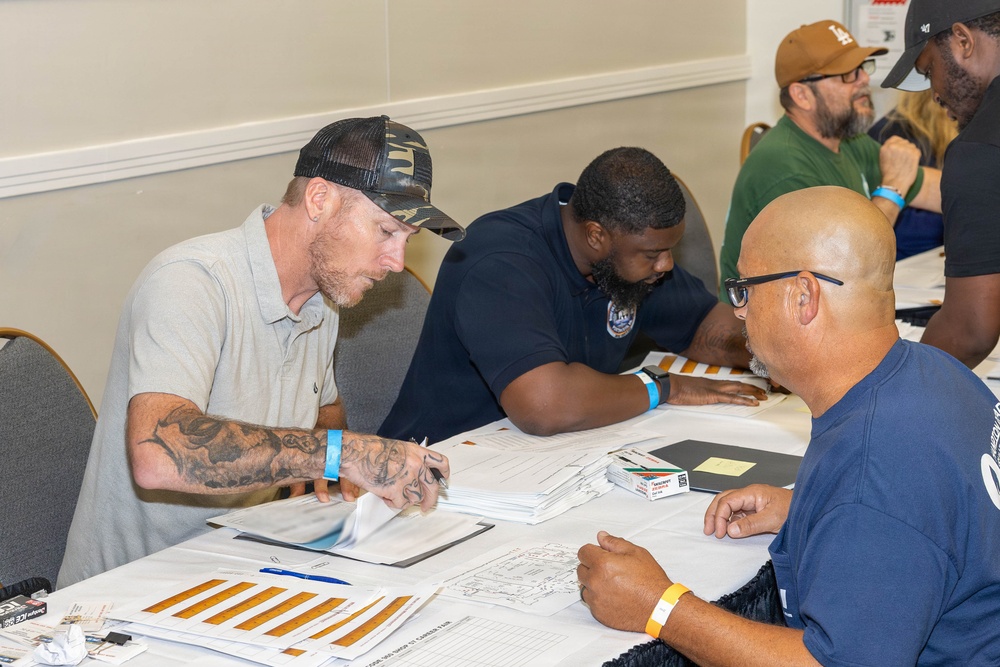 A group of men are gathered around a table in what appears to be a career fair or registration event, reviewing and filling out paperwork. Several participants are wearing colored wristbands, suggesting an organized event with attendee tracking. The setting appears to be a conference or community center room with multiple tables in use.