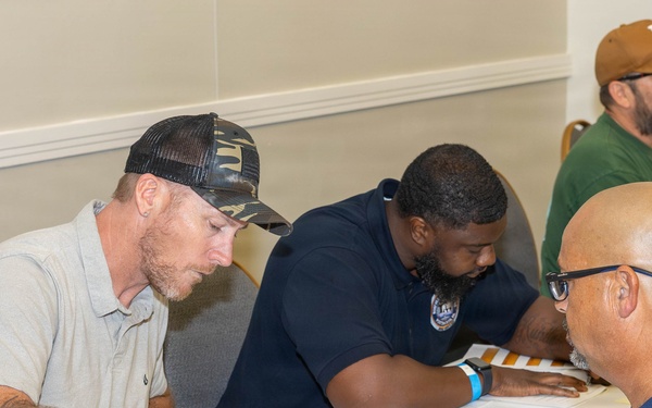 A group of men are gathered around a table in what appears to be a career fair or registration event, reviewing and filling out paperwork. Several participants are wearing colored wristbands, suggesting an organized event with attendee tracking. The setting appears to be a conference or community center room with multiple tables in use.