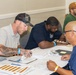 A group of men are gathered around a table in what appears to be a career fair or registration event, reviewing and filling out paperwork. Several participants are wearing colored wristbands, suggesting an organized event with attendee tracking. The setting appears to be a conference or community center room with multiple tables in use.