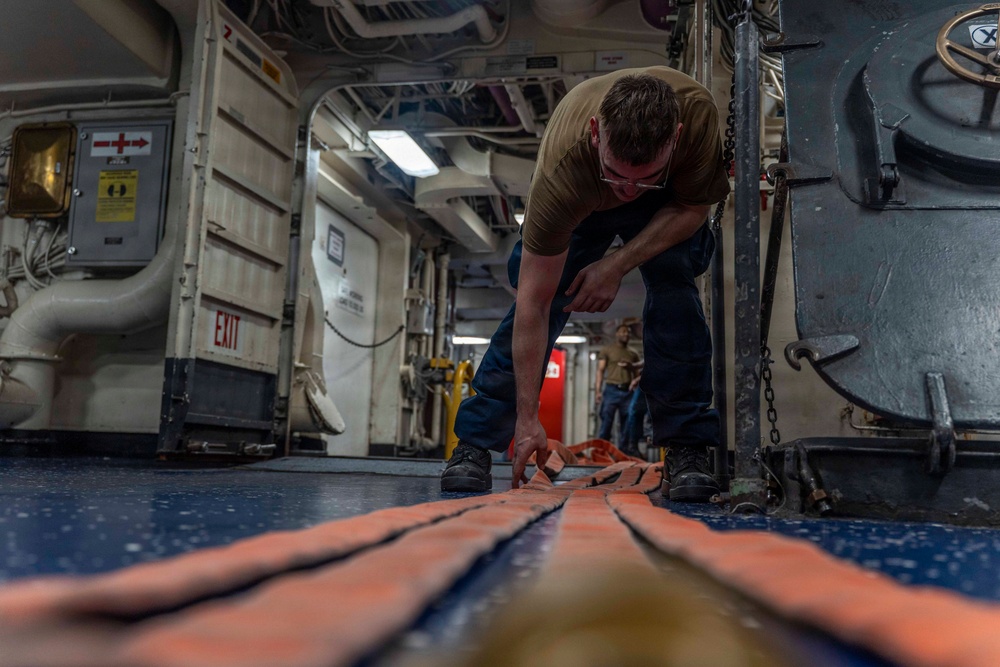 A sailor or military personnel is crouched down on the deck of what appears to be a naval vessel, working with orange tie-down straps or securing equipment on the floor. The interior space shows typical naval ship characteristics including pipes, conduits, safety signage, and industrial equipment. A second person is visible in the background of the corridor.