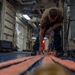 A sailor or military personnel is crouched down on the deck of what appears to be a naval vessel, working with orange tie-down straps or securing equipment on the floor. The interior space shows typical naval ship characteristics including pipes, conduits, safety signage, and industrial equipment. A second person is visible in the background of the corridor.