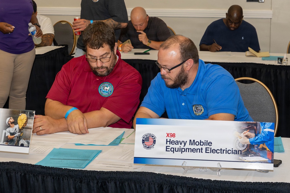 Two men wearing polo shirts with naval shipyard emblems are seated at a table reviewing documents at what appears to be a professional meeting or job fair. A nameplate in front of them reads 'X98 Heavy Mobile Equipment Electrician' from America's Naval Shipyards. Several other participants are visible in the background, also engaged in paperwork or discussion.