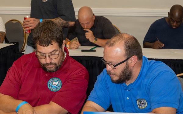 Two men wearing polo shirts with naval shipyard emblems are seated at a table reviewing documents at what appears to be a professional meeting or job fair. A nameplate in front of them reads 'X98 Heavy Mobile Equipment Electrician' from America's Naval Shipyards. Several other participants are visible in the background, also engaged in paperwork or discussion.