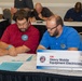 Two men wearing polo shirts with naval shipyard emblems are seated at a table reviewing documents at what appears to be a professional meeting or job fair. A nameplate in front of them reads 'X98 Heavy Mobile Equipment Electrician' from America's Naval Shipyards. Several other participants are visible in the background, also engaged in paperwork or discussion.