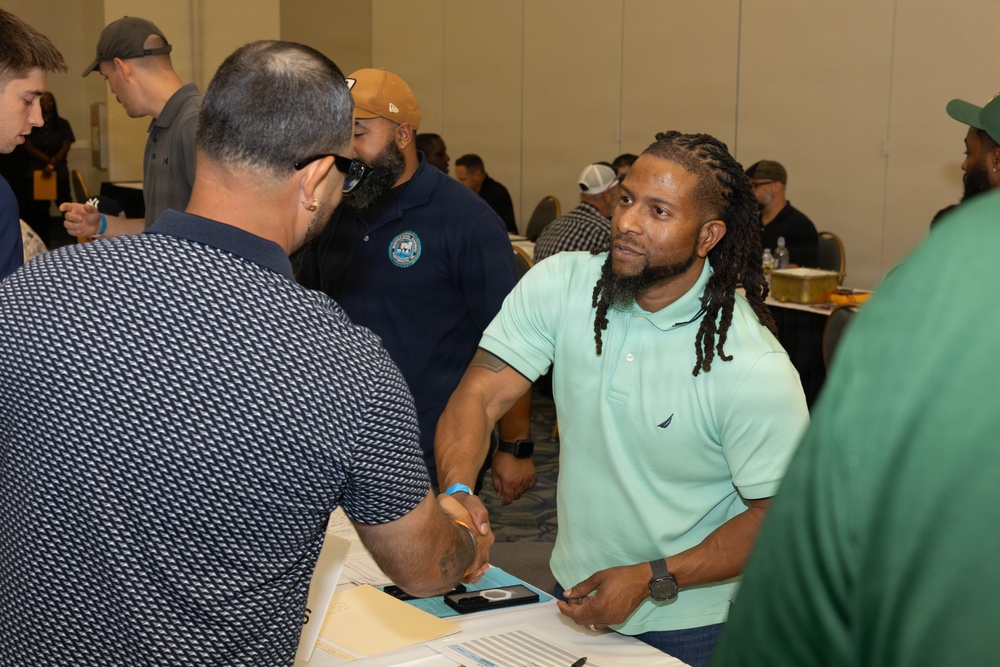 The image depicts a professional networking or job fair event taking place in a conference or banquet hall. Two men are prominently shown shaking hands across a table covered with documents and papers. The room is filled with numerous other attendees engaged in similar interactions in the background.