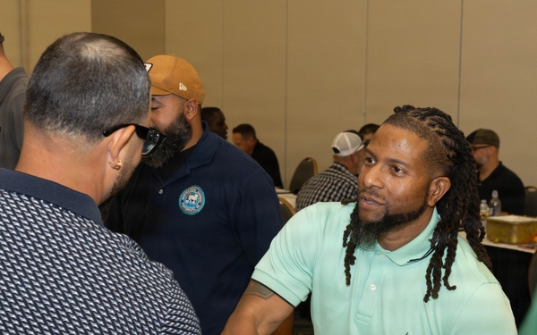 The image depicts a professional networking or job fair event taking place in a conference or banquet hall. Two men are prominently shown shaking hands across a table covered with documents and papers. The room is filled with numerous other attendees engaged in similar interactions in the background.