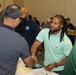 The image depicts a professional networking or job fair event taking place in a conference or banquet hall. Two men are prominently shown shaking hands across a table covered with documents and papers. The room is filled with numerous other attendees engaged in similar interactions in the background.