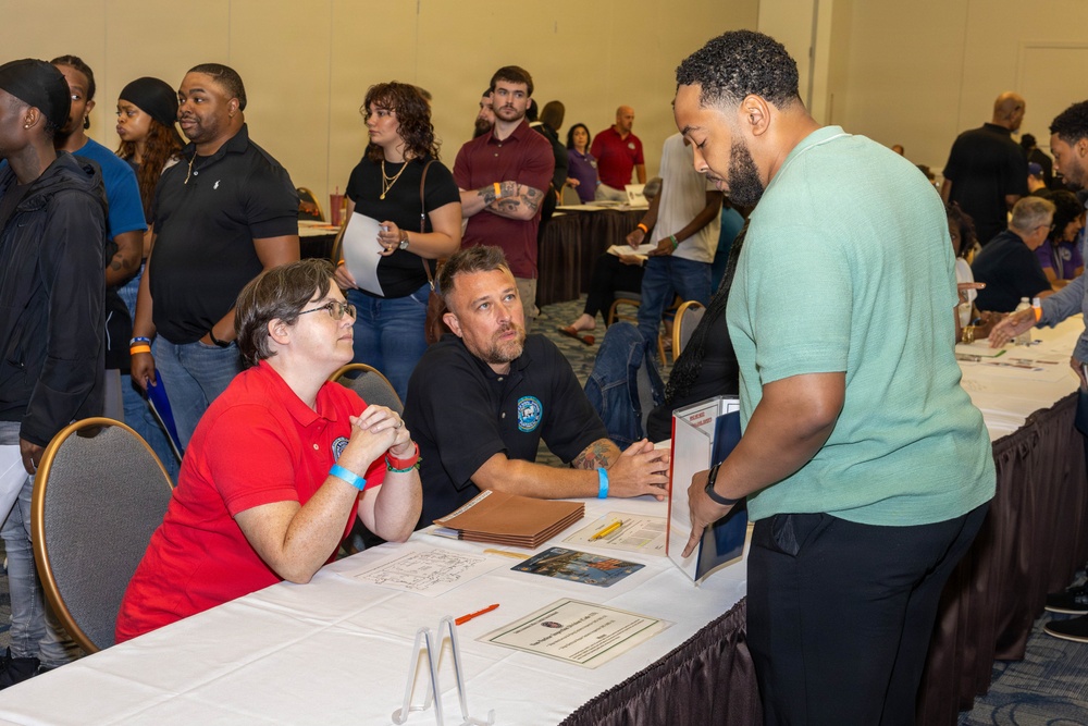 The image depicts what appears to be a job fair or recruitment event held in a large conference or banquet hall. Two representatives seated at a table with informational materials are engaged in conversation with a standing attendee. The venue is crowded with numerous other participants visible in the background.
