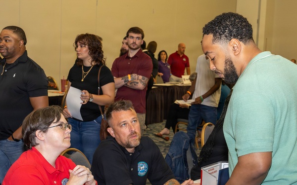 The image depicts what appears to be a job fair or recruitment event held in a large conference or banquet hall. Two representatives seated at a table with informational materials are engaged in conversation with a standing attendee. The venue is crowded with numerous other participants visible in the background.