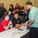 The image depicts what appears to be a job fair or recruitment event held in a large conference or banquet hall. Two representatives seated at a table with informational materials are engaged in conversation with a standing attendee. The venue is crowded with numerous other participants visible in the background.