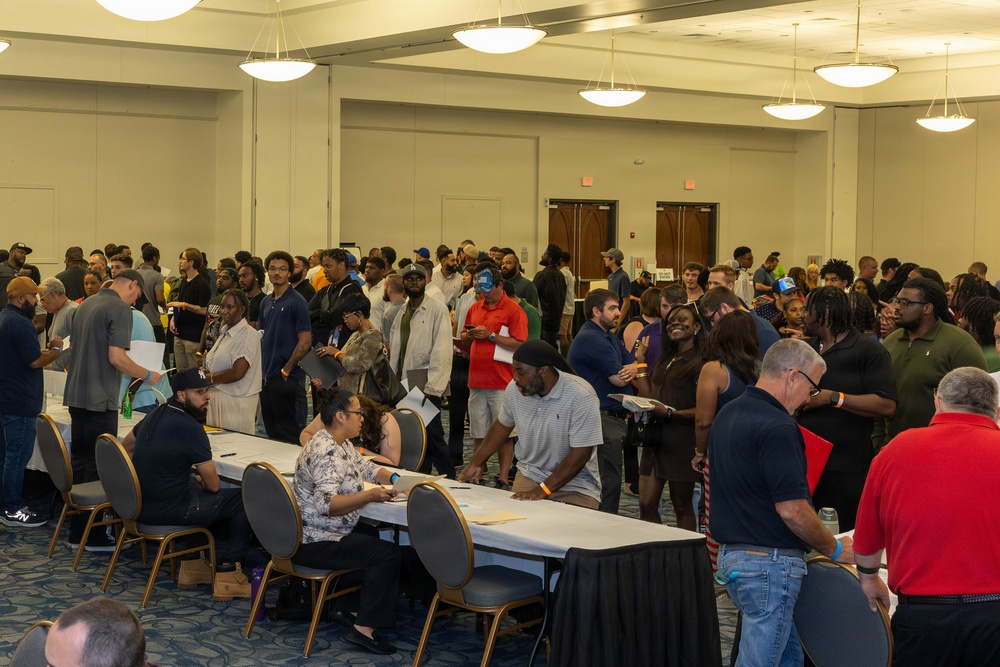 A large indoor gathering in what appears to be a convention center or hotel ballroom, with a long registration or check-in table staffed by seated individuals processing a crowd of attendees. The event draws a diverse group of people, many holding papers or documents, standing in lines or clusters throughout the room. The setting and activity suggest a job fair, community event, or organized registration process.