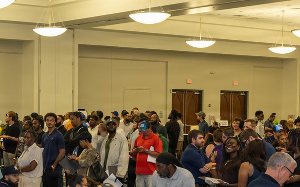 A large indoor gathering in what appears to be a convention center or hotel ballroom, with a long registration or check-in table staffed by seated individuals processing a crowd of attendees. The event draws a diverse group of people, many holding papers or documents, standing in lines or clusters throughout the room. The setting and activity suggest a job fair, community event, or organized registration process.