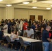 A large indoor gathering in what appears to be a convention center or hotel ballroom, with a long registration or check-in table staffed by seated individuals processing a crowd of attendees. The event draws a diverse group of people, many holding papers or documents, standing in lines or clusters throughout the room. The setting and activity suggest a job fair, community event, or organized registration process.