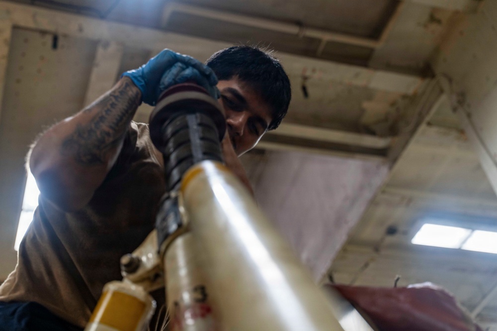 A young male technician wearing blue nitrile gloves and a face mask is working on a large mechanical component, likely part of an aircraft or heavy machinery. He appears to be inspecting or assembling a cylindrical assembly with black rubber seals and yellow-painted metal tubing. The photo is taken from a low angle inside an industrial or hangar-type facility.