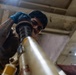 A young male technician wearing blue nitrile gloves and a face mask is working on a large mechanical component, likely part of an aircraft or heavy machinery. He appears to be inspecting or assembling a cylindrical assembly with black rubber seals and yellow-painted metal tubing. The photo is taken from a low angle inside an industrial or hangar-type facility.