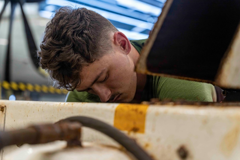 A young male technician or mechanic is closely inspecting or working on a piece of heavy equipment or machinery. He is leaning in with his head down, focused on an area between metal panels or compartments. The setting appears to be an industrial workshop or maintenance bay.
