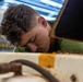 A young male technician or mechanic is closely inspecting or working on a piece of heavy equipment or machinery. He is leaning in with his head down, focused on an area between metal panels or compartments. The setting appears to be an industrial workshop or maintenance bay.