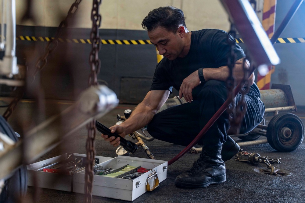 A man dressed in all-black clothing and boots is crouching on a dark floor in what appears to be an industrial or hangar-type facility. He is handling a hose or pneumatic tool connected to a valve assembly, with an open white tool case containing organized tools in front of him. The environment includes heavy chains, wheeled equipment, and yellow-black hazard markings on walls in the background.