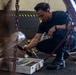 A man dressed in all-black clothing and boots is crouching on a dark floor in what appears to be an industrial or hangar-type facility. He is handling a hose or pneumatic tool connected to a valve assembly, with an open white tool case containing organized tools in front of him. The environment includes heavy chains, wheeled equipment, and yellow-black hazard markings on walls in the background.