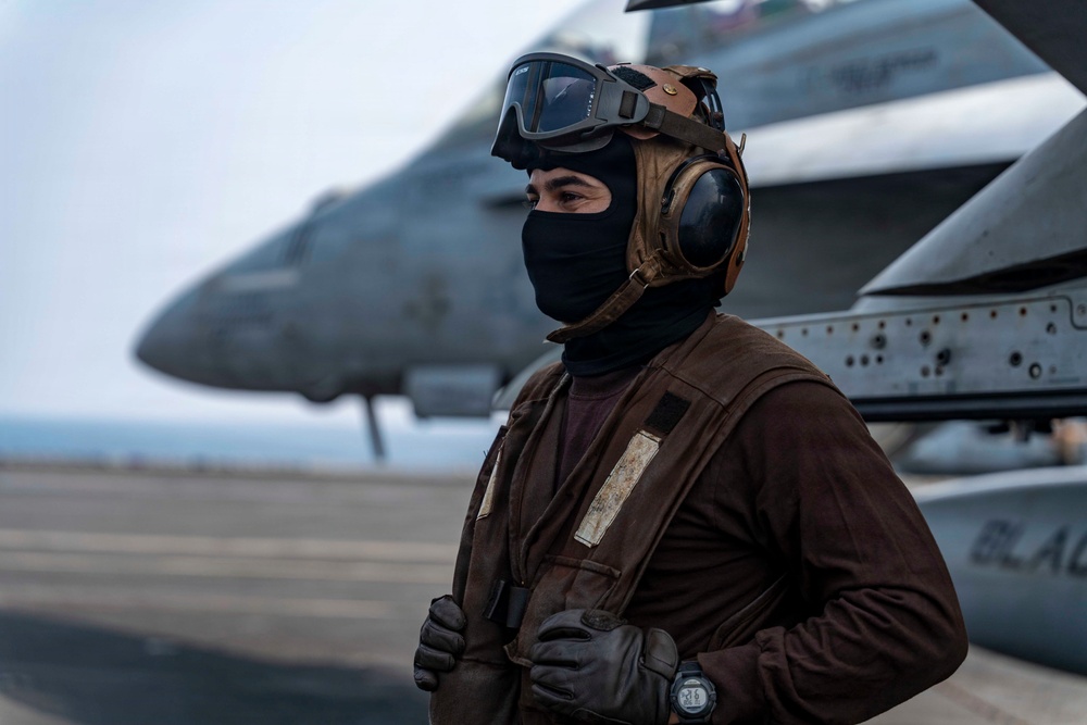 A military flight deck crew member stands on an aircraft carrier flight deck wearing full protective gear including a brown jersey, helmet with ear protection, goggles, and a black balaclava. Military jet aircraft, consistent with F/A-18 Hornets or Super Hornets, are visible in the background on the flight deck. The brown jersey color indicates the individual is likely an aircraft handler or plane captain in the U.S. Navy.