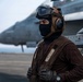 A military flight deck crew member stands on an aircraft carrier flight deck wearing full protective gear including a brown jersey, helmet with ear protection, goggles, and a black balaclava. Military jet aircraft, consistent with F/A-18 Hornets or Super Hornets, are visible in the background on the flight deck. The brown jersey color indicates the individual is likely an aircraft handler or plane captain in the U.S. Navy.