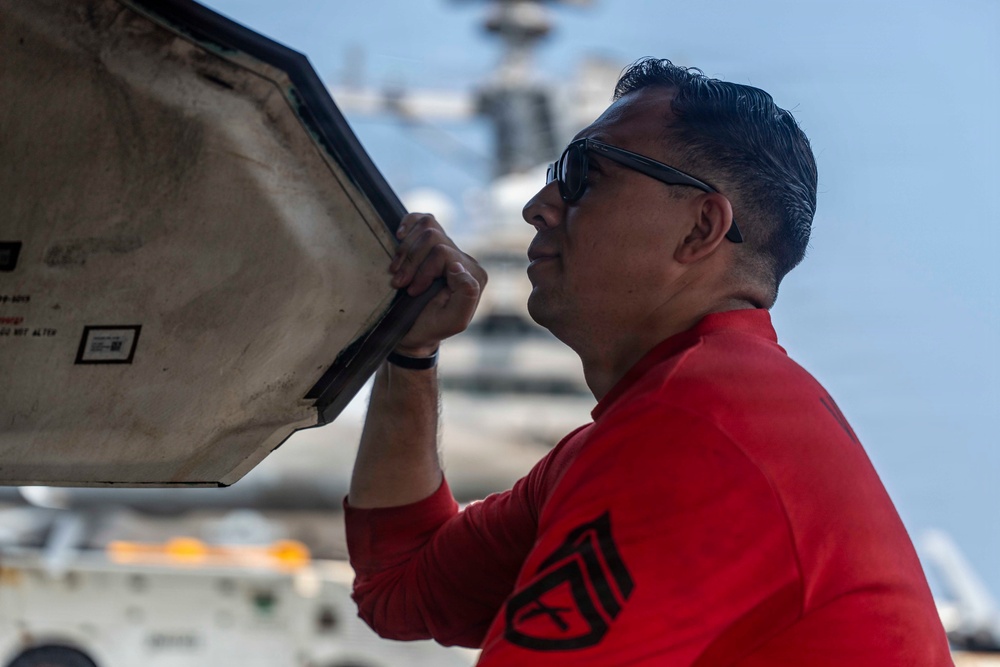 A man wearing a red long-sleeve shirt and sunglasses is inspecting or opening an access panel on what appears to be a military aircraft. The setting appears to be the flight deck of an aircraft carrier, with additional naval vessels visible in the background. The image is a close-up profile shot with shallow depth of field.