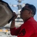 A man wearing a red long-sleeve shirt and sunglasses is inspecting or opening an access panel on what appears to be a military aircraft. The setting appears to be the flight deck of an aircraft carrier, with additional naval vessels visible in the background. The image is a close-up profile shot with shallow depth of field.