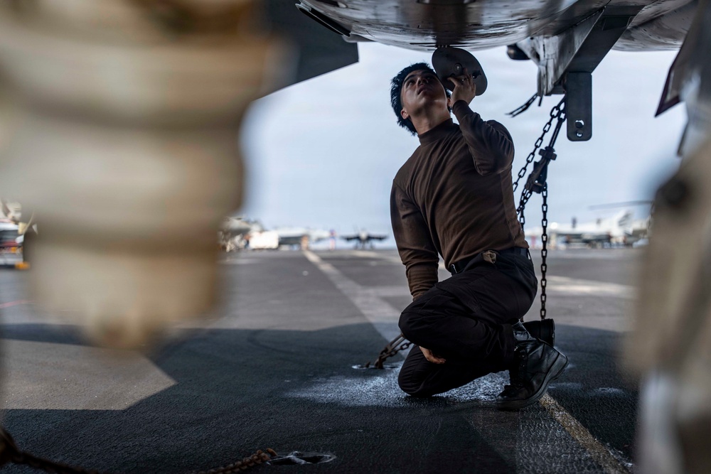 A military technician or aviation mechanic is kneeling beneath an aircraft on what appears to be a naval aircraft carrier flight deck. The individual is performing maintenance or inspection work on the underside of the aircraft, handling a component near a tie-down chain. Additional aircraft and deck equipment are visible in the background.