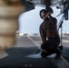 A military technician or aviation mechanic is kneeling beneath an aircraft on what appears to be a naval aircraft carrier flight deck. The individual is performing maintenance or inspection work on the underside of the aircraft, handling a component near a tie-down chain. Additional aircraft and deck equipment are visible in the background.