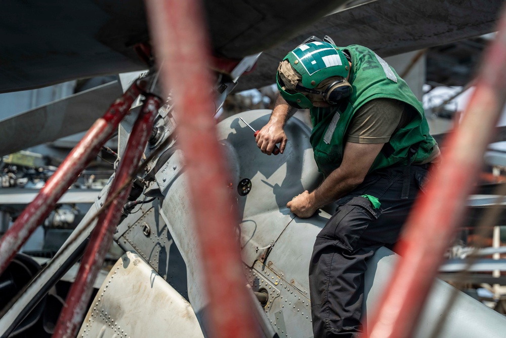 A military aviation technician wearing a green jersey and helmet with hearing protection is performing maintenance work on a military aircraft, likely on the deck of an aircraft carrier. The worker is leaning over the aircraft fuselage and appears to be using a hand tool. The image is shot through red safety barriers or tie-down equipment in the foreground, creating a framed perspective.