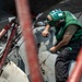 A military aviation technician wearing a green jersey and helmet with hearing protection is performing maintenance work on a military aircraft, likely on the deck of an aircraft carrier. The worker is leaning over the aircraft fuselage and appears to be using a hand tool. The image is shot through red safety barriers or tie-down equipment in the foreground, creating a framed perspective.