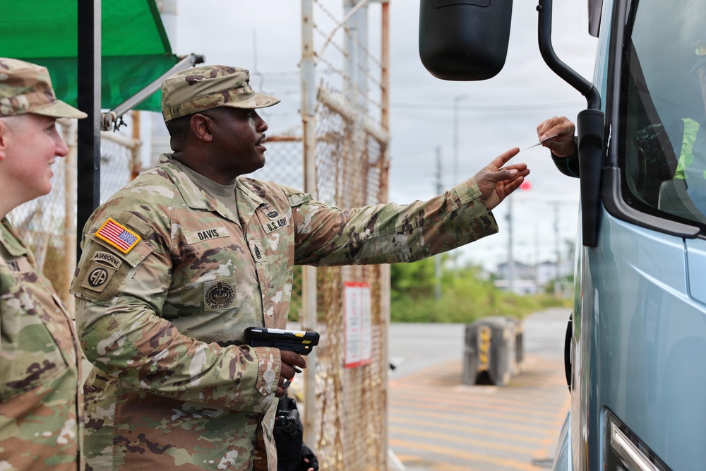 A U.S. Army soldier named Davis, wearing OCP (Operational Camouflage Pattern) uniform with 82nd Airborne Division patch, is handing or receiving a document at what appears to be a military installation gate checkpoint. A second soldier is visible in the background to the left. The scene depicts a routine access control or vehicle inspection procedure at a secured entry point.
