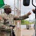 A U.S. Army soldier named Davis, wearing OCP (Operational Camouflage Pattern) uniform with 82nd Airborne Division patch, is handing or receiving a document at what appears to be a military installation gate checkpoint. A second soldier is visible in the background to the left. The scene depicts a routine access control or vehicle inspection procedure at a secured entry point.