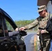 A U.S. Army soldier in MultiCam/OCP camouflage uniform is conducting a vehicle checkpoint inspection, handing or receiving a document from a vehicle occupant. A second individual wearing a tactical vest and civilian clothing stands nearby holding a handheld scanning device. The scene appears to take place at a military installation entry control point.