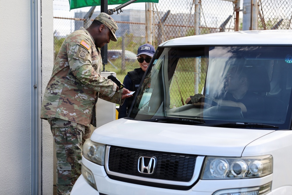 A U.S. Army soldier in OCP (Operational Camouflage Pattern) uniform is conducting a vehicle checkpoint inspection at what appears to be a military installation gate. He is checking documents from a driver wearing a 'SECURITY' cap seated in a white Honda Element. The interaction appears routine and friendly, with the driver smiling.