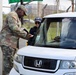 A U.S. Army soldier in OCP (Operational Camouflage Pattern) uniform is conducting a vehicle checkpoint inspection at what appears to be a military installation gate. He is checking documents from a driver wearing a 'SECURITY' cap seated in a white Honda Element. The interaction appears routine and friendly, with the driver smiling.