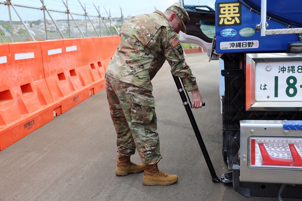 A U.S. Army soldier in MultiCam (OCP) camouflage uniform is using an inspection mirror or pole tool to examine the underside of a Japanese commercial truck. The setting appears to be a military checkpoint or gate in Okinawa, Japan, indicated by the Japanese license plate and signage on the vehicle. Orange plastic barriers (YODOCK brand) line the inspection lane.