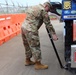 A U.S. Army soldier in MultiCam (OCP) camouflage uniform is using an inspection mirror or pole tool to examine the underside of a Japanese commercial truck. The setting appears to be a military checkpoint or gate in Okinawa, Japan, indicated by the Japanese license plate and signage on the vehicle. Orange plastic barriers (YODOCK brand) line the inspection lane.