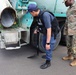 A security officer wearing a tactical vest labeled 'SECURITY' is inspecting the underside of a cement mixer truck using a telescoping mirror inspection tool. A U.S. Army soldier in OCP (Operational Camouflage Pattern) uniform stands nearby observing the inspection. This appears to be a vehicle security checkpoint inspection, likely at a military installation.