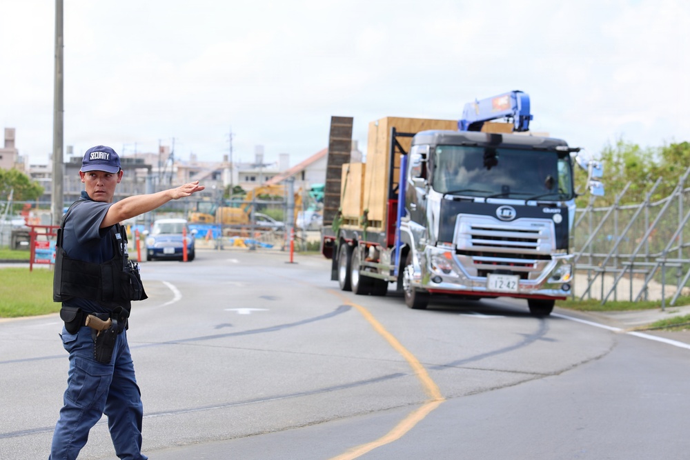 A security officer wearing a blue uniform, vest, and cap labeled 'SECURITY' is directing traffic at what appears to be a construction site entrance. A large flatbed truck carrying wooden crates or construction materials is approaching, with a crane attachment visible on top. The scene appears to be in Japan, based on the Japanese license plate and vehicle styling.