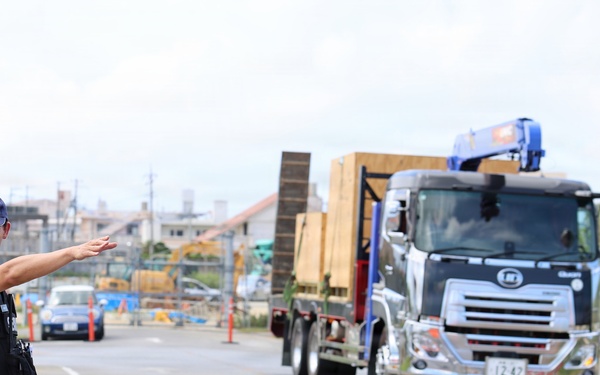 A security officer wearing a blue uniform, vest, and cap labeled 'SECURITY' is directing traffic at what appears to be a construction site entrance. A large flatbed truck carrying wooden crates or construction materials is approaching, with a crane attachment visible on top. The scene appears to be in Japan, based on the Japanese license plate and vehicle styling.