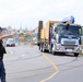 A security officer wearing a blue uniform, vest, and cap labeled 'SECURITY' is directing traffic at what appears to be a construction site entrance. A large flatbed truck carrying wooden crates or construction materials is approaching, with a crane attachment visible on top. The scene appears to be in Japan, based on the Japanese license plate and vehicle styling.