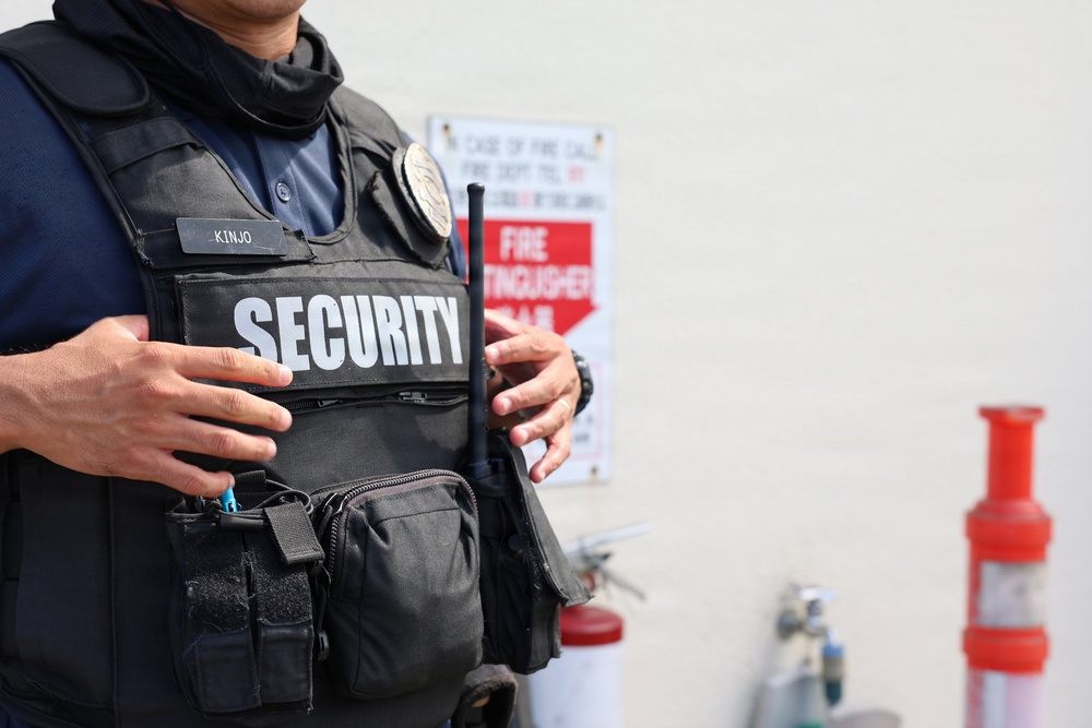 The image shows the torso of a security guard wearing a black tactical vest with 'SECURITY' printed in large white letters on the front. A name tag reading 'KINJO' is visible on the vest, along with a badge and a radio/walkie-talkie. In the background, fire extinguishers and a fire extinguisher location sign are visible against a light-colored wall.