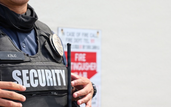 The image shows the torso of a security guard wearing a black tactical vest with 'SECURITY' printed in large white letters on the front. A name tag reading 'KINJO' is visible on the vest, along with a badge and a radio/walkie-talkie. In the background, fire extinguishers and a fire extinguisher location sign are visible against a light-colored wall.