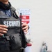 The image shows the torso of a security guard wearing a black tactical vest with 'SECURITY' printed in large white letters on the front. A name tag reading 'KINJO' is visible on the vest, along with a badge and a radio/walkie-talkie. In the background, fire extinguishers and a fire extinguisher location sign are visible against a light-colored wall.