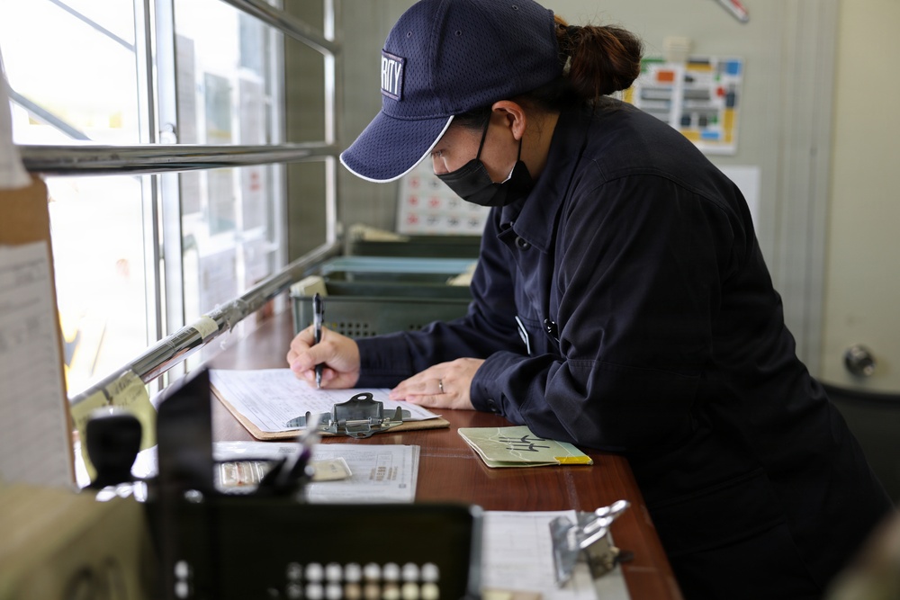 A security personnel wearing a dark navy uniform and cap with partial text 'RITY' (likely 'SECURITY') is writing on a clipboard at a desk or counter station. The individual is wearing a black face mask and appears to be filling out paperwork or a log form. The setting appears to be a security checkpoint or guard station with a service window.