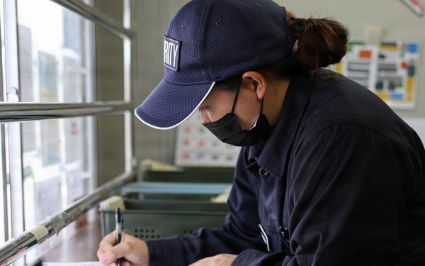 A security personnel wearing a dark navy uniform and cap with partial text 'RITY' (likely 'SECURITY') is writing on a clipboard at a desk or counter station. The individual is wearing a black face mask and appears to be filling out paperwork or a log form. The setting appears to be a security checkpoint or guard station with a service window.