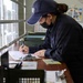 A security personnel wearing a dark navy uniform and cap with partial text 'RITY' (likely 'SECURITY') is writing on a clipboard at a desk or counter station. The individual is wearing a black face mask and appears to be filling out paperwork or a log form. The setting appears to be a security checkpoint or guard station with a service window.