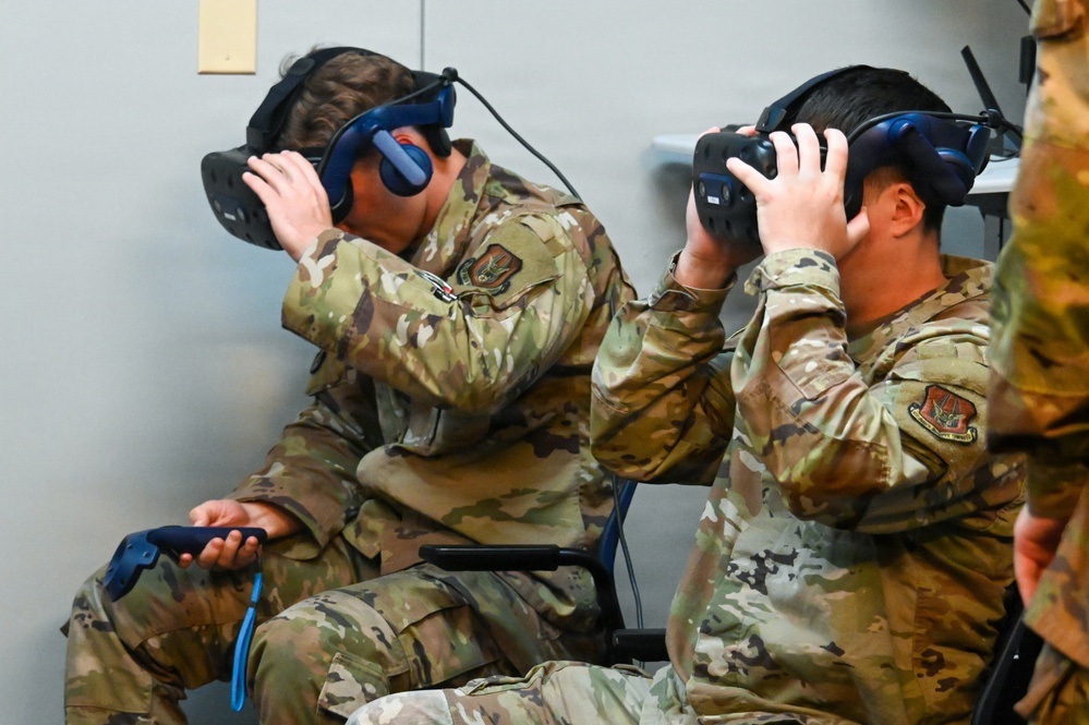 Two military personnel in OCP (Operational Camouflage Pattern) uniforms are seated and using virtual reality headsets, holding VR controllers. Both individuals are adjusting or wearing their VR headsets simultaneously, suggesting a training or demonstration session. A third uniformed individual is partially visible on the right edge of the frame.
