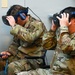 Two military personnel in OCP (Operational Camouflage Pattern) uniforms are seated and using virtual reality headsets, holding VR controllers. Both individuals are adjusting or wearing their VR headsets simultaneously, suggesting a training or demonstration session. A third uniformed individual is partially visible on the right edge of the frame.